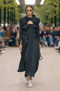 Model walking a fashion show in a dark, structured coat-dress with gold buttons and white sneakers, holding a small bag.