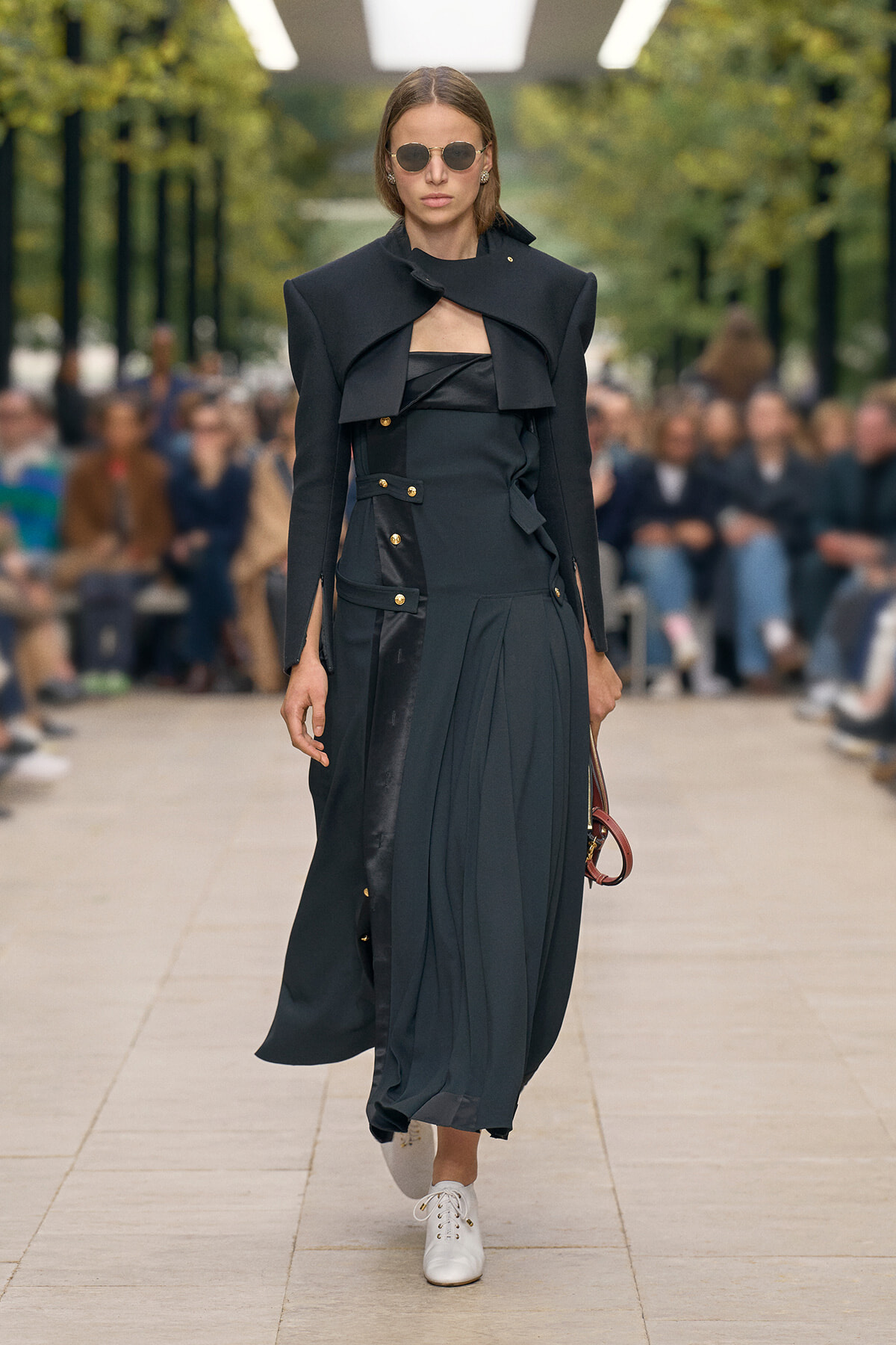 Model walking a fashion show in a dark, structured coat-dress with gold buttons and white sneakers, holding a small bag.