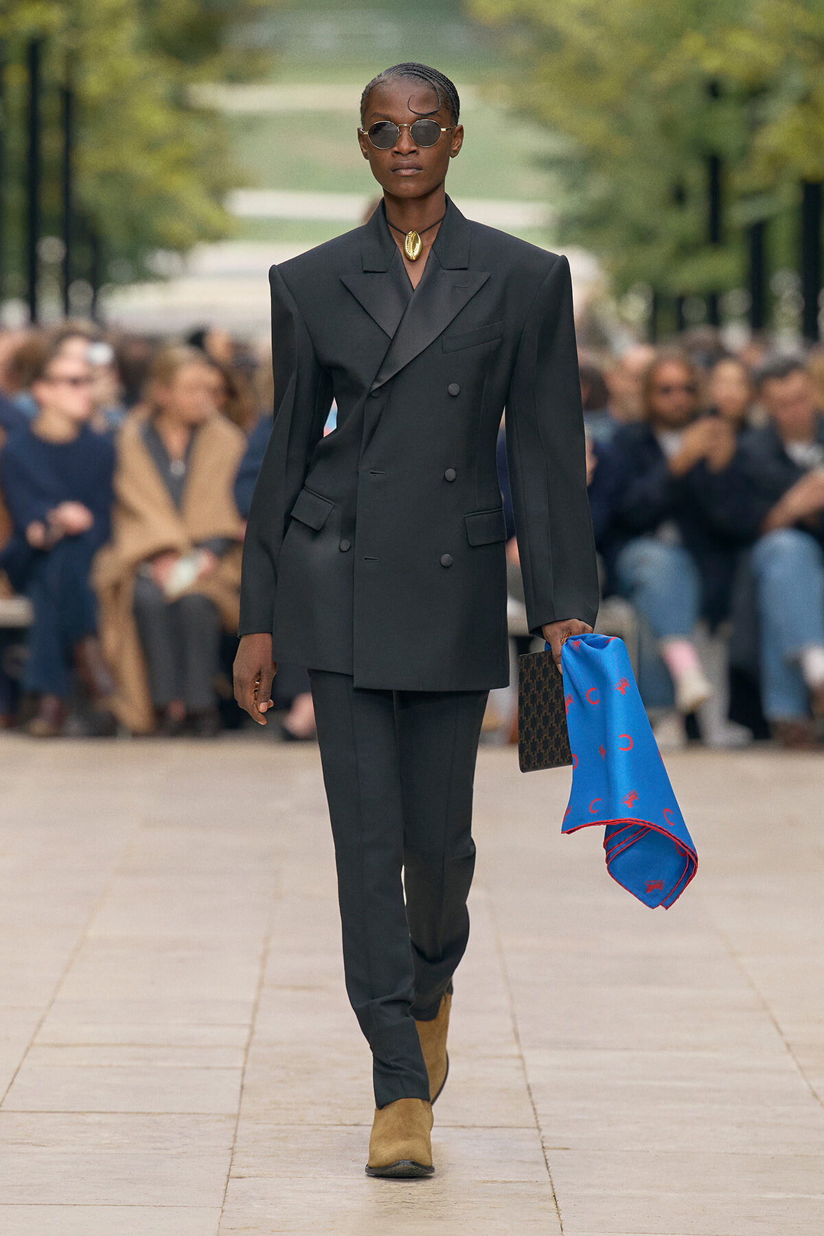 Model walks a runway in an oversized black double-breasted blazer, matching trousers, tan boots, sunglasses, and a bright blue scarf in her hand.