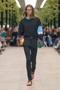 Full-length shot of a female model walking a runway in a black outfit: loose black top with multicolor sleeves, black pants, belt, and brown sandals, audience in background.