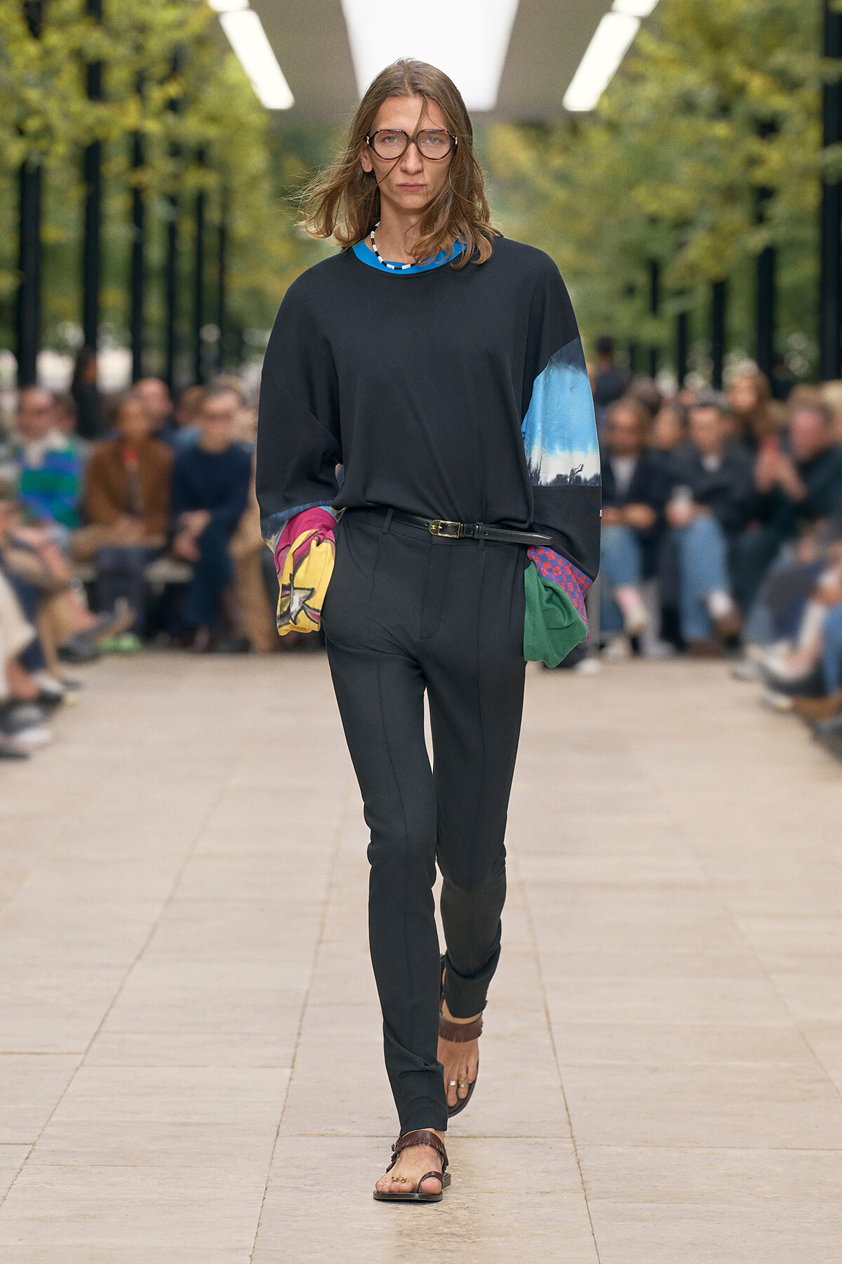 Full-length shot of a female model walking a runway in a black outfit: loose black top with multicolor sleeves, black pants, belt, and brown sandals, audience in background.