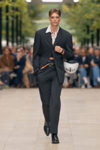 Male model in a black suit walks a runway, holding a white helmet labeled CELINE PARIS.