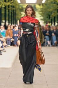 Model walking a fashion runway outdoors in a bold, patterned top with a red scarf, black wide-leg pants, and a brown handbag.