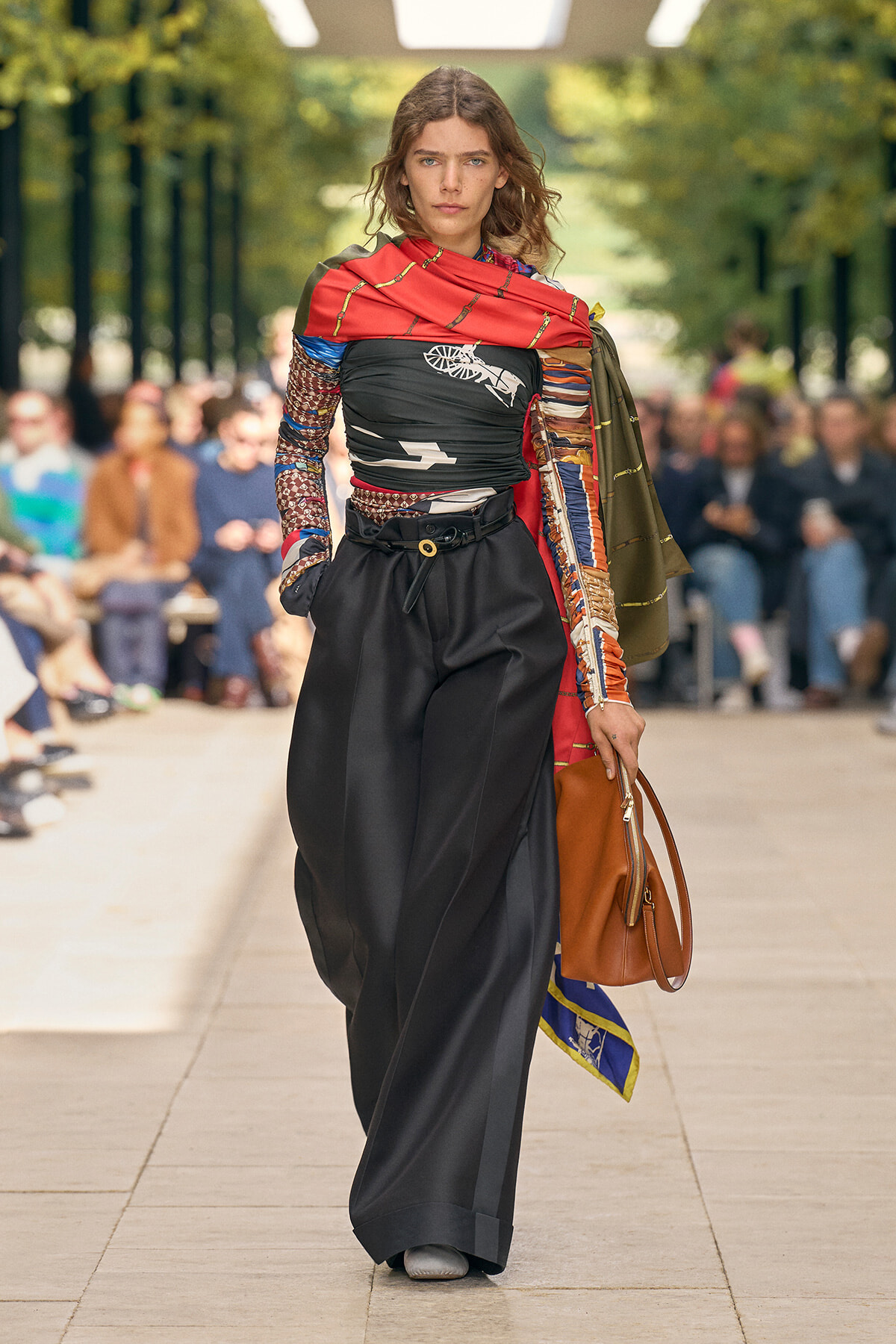 Model walking a fashion runway outdoors in a bold, patterned top with a red scarf, black wide-leg pants, and a brown handbag.