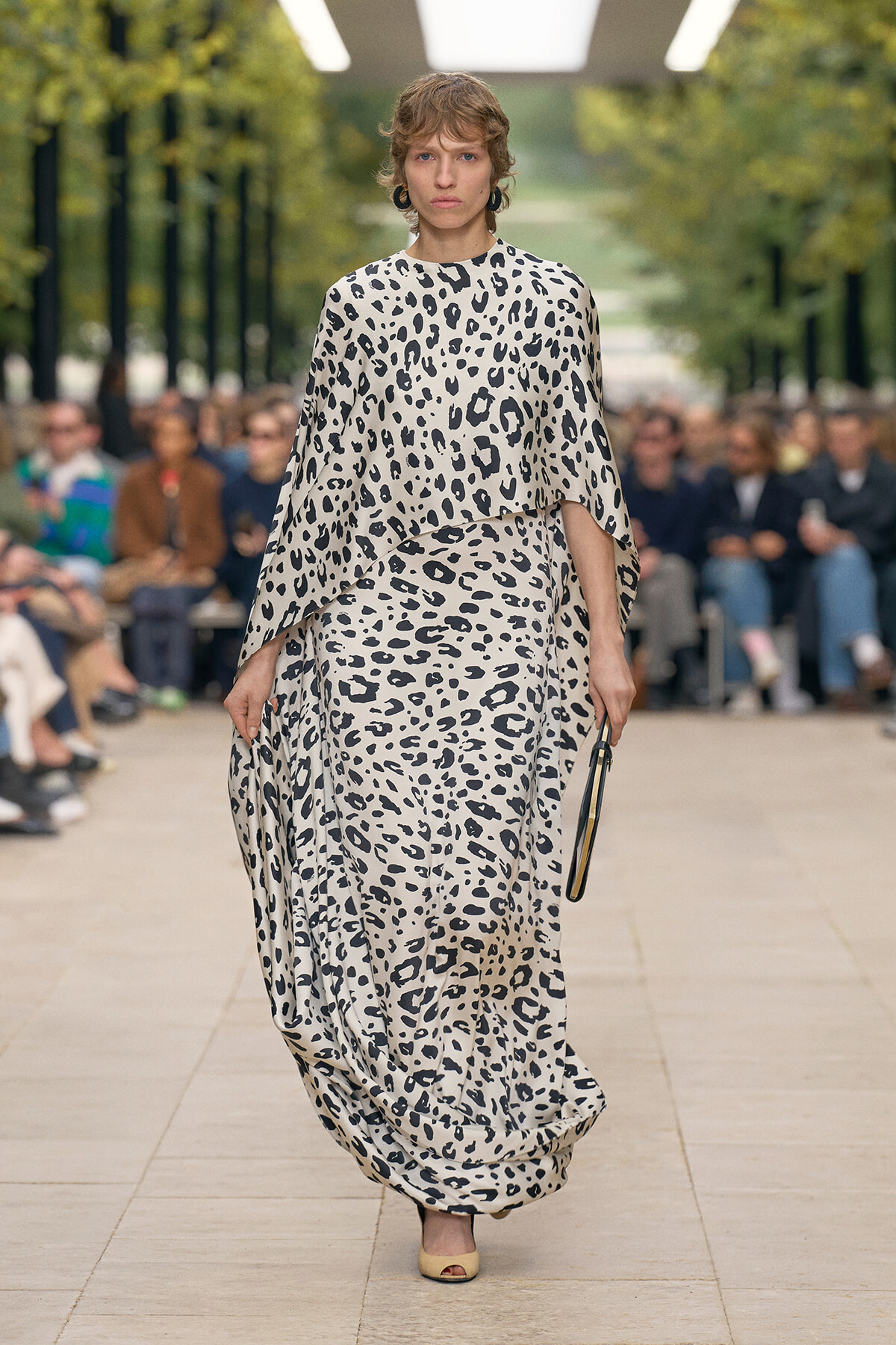 models walks a monochrome leopard-print flowing gown down a runway in an outdoor fashion show.
