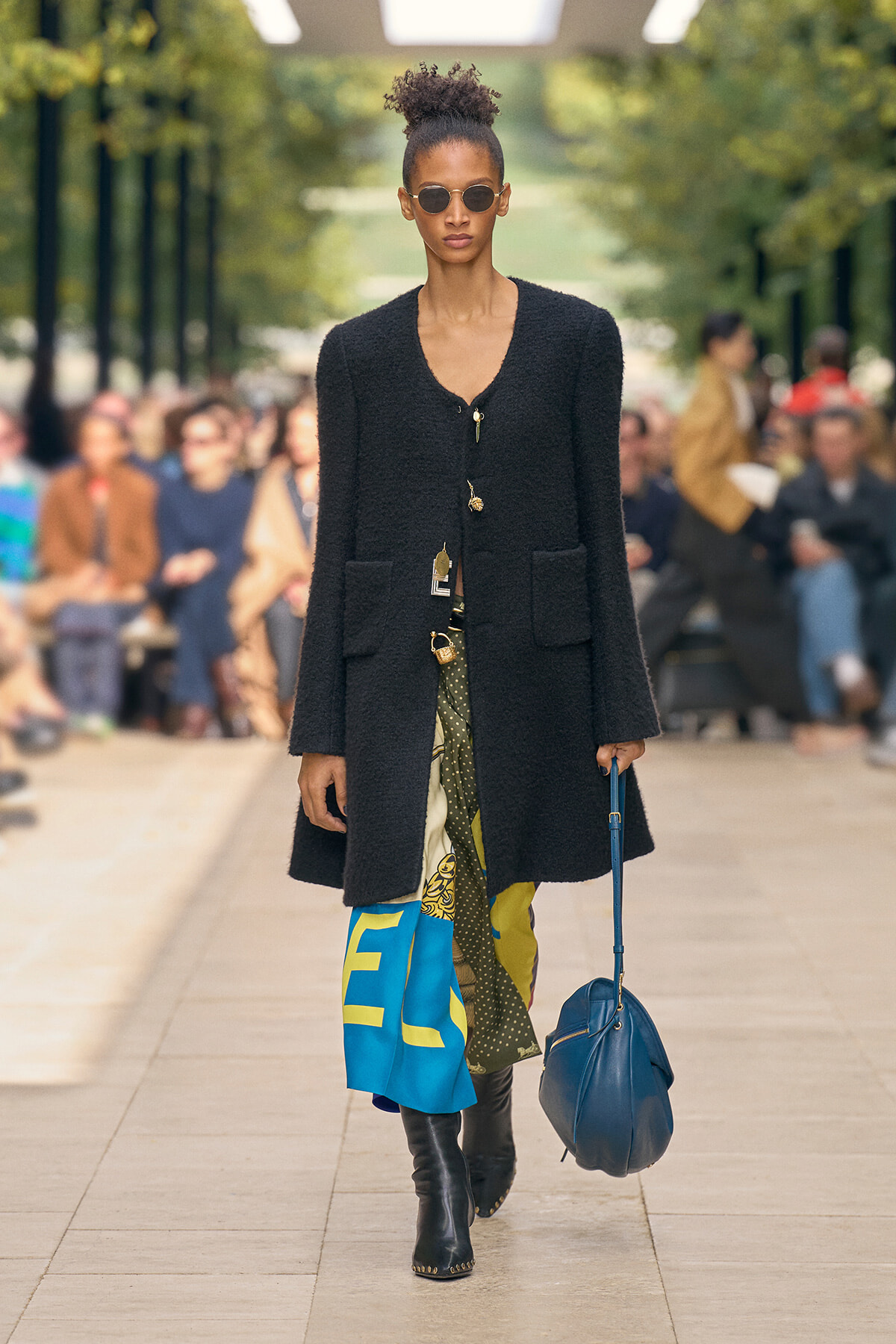 Model walks a runway in a black textured coat with large pockets, colorful patterned pants, black boots, round sunglasses, and a blue handbag.