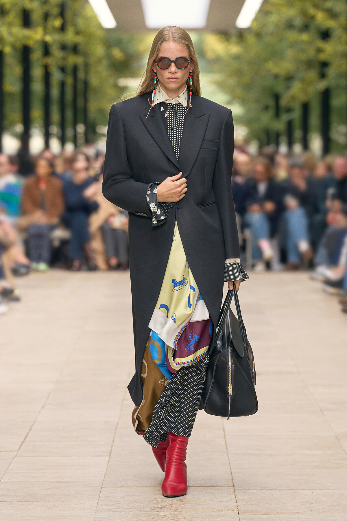 Model walking a runway in a long black blazer over a polka-dot blouse, colorful scarf peeking from the slit, red boots, round sunglasses, and a large black bag; audience blurred in the background.
