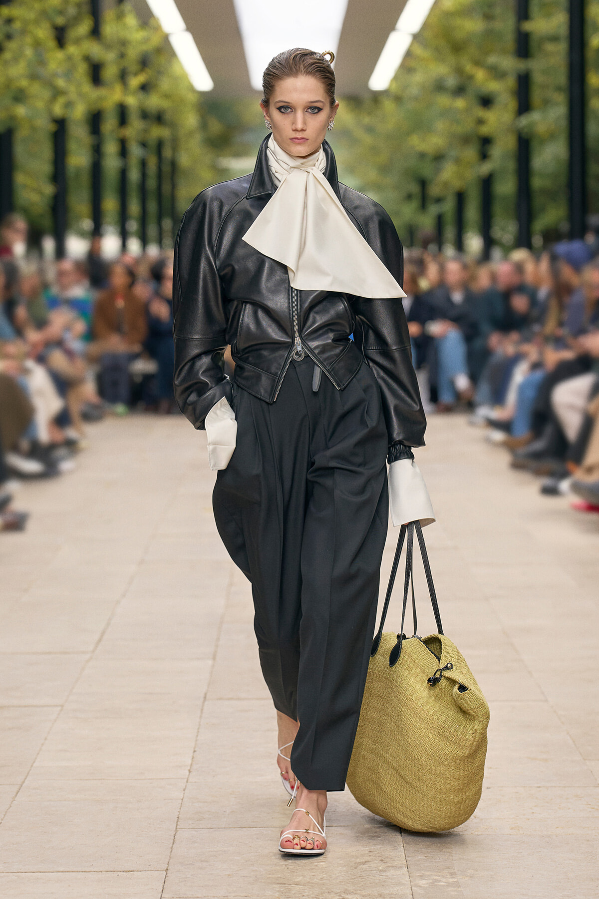 Model walks a runway in a black leather jacket with a large cream scarf, black wide-leg trousers, white cuffs, and a large woven straw tote.