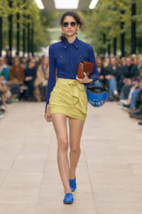 Model walks a runway in a cobalt blue button-down blouse and pale yellow wrapped mini-skirt, with blue shoes, sunglasses, and a brown crocodile-textured clutch plus a blue helmet.