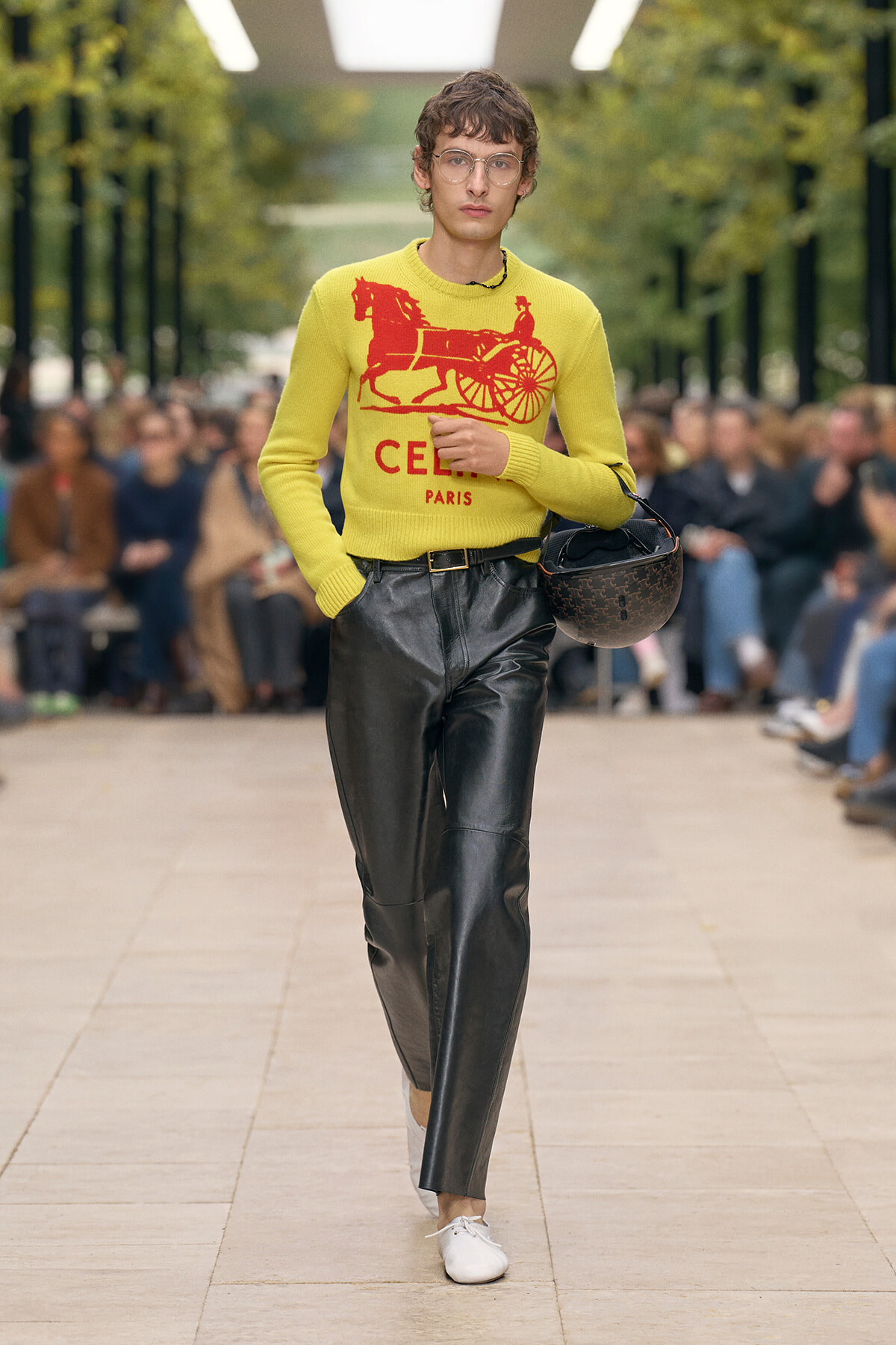 Model walks a fashion show in a bright yellow sweater with a red horse-drawn carriage graphic and 'CEL PARIS', paired with black leather pants and white shoes, holding a brown helmet.