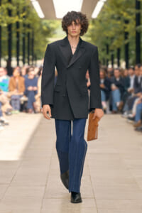 Male model on a fashion runway wearing an oversized black double‑breasted blazer, blue jeans, black shoes, and holding a brown portfolio; audience in background.