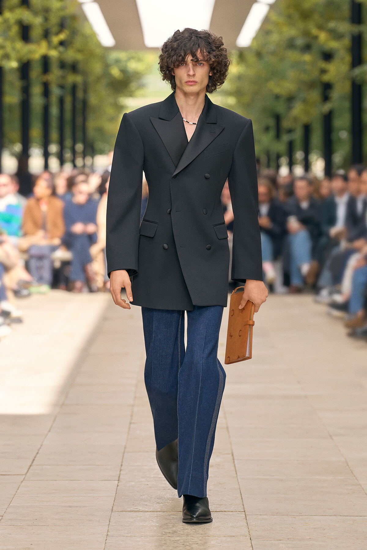 Male model on a fashion runway wearing an oversized black double‑breasted blazer, blue jeans, black shoes, and holding a brown portfolio; audience in background.