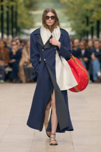 Model walking a runway in a navy double-breasted trench with a cream scarf and large red tote bag, wearing round sunglasses.