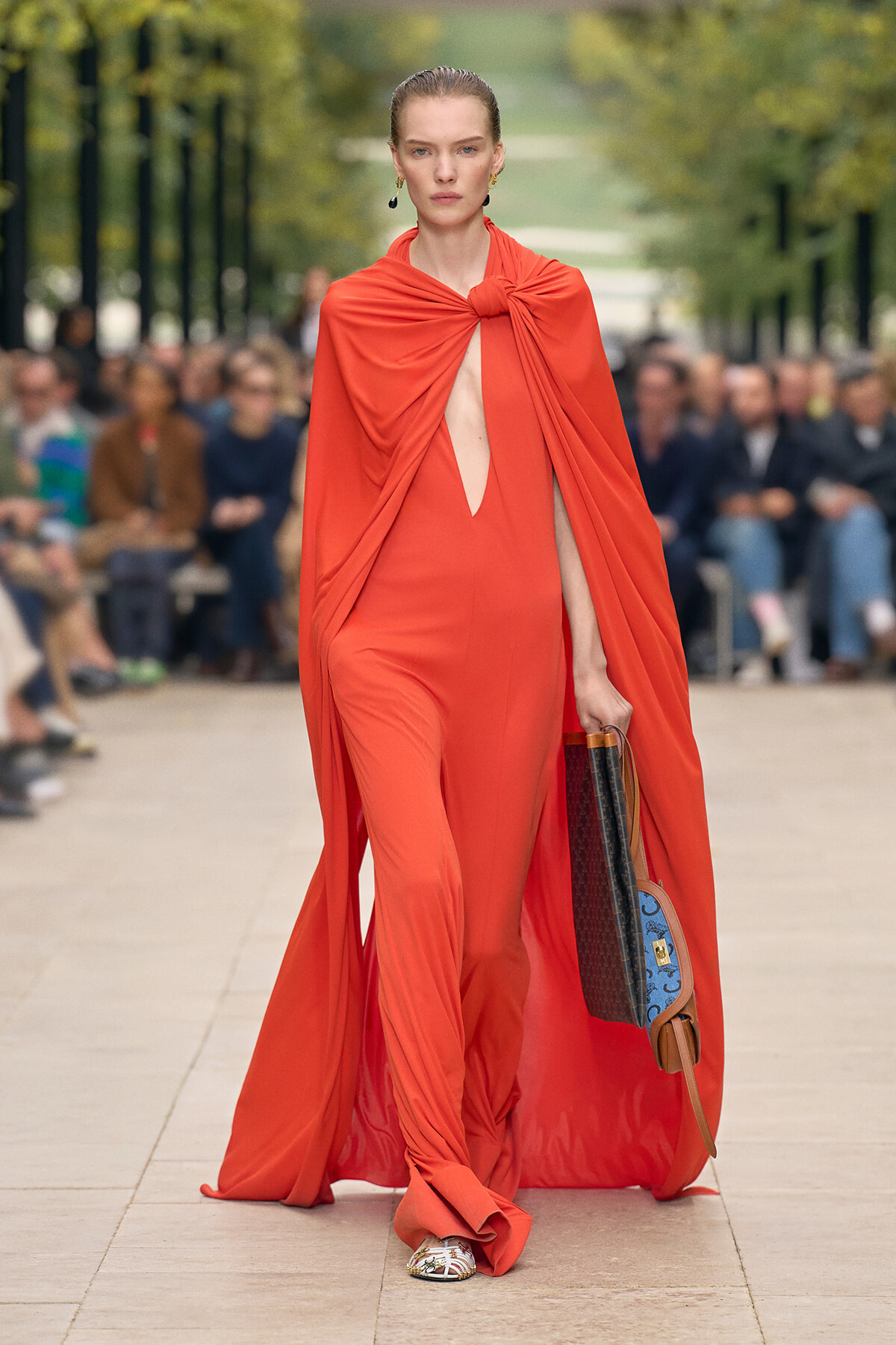 Model walks a runway in a vivid orange cape-style gown with a knot at the neckline and a slitted bodice, carrying a brown/blue handbag.