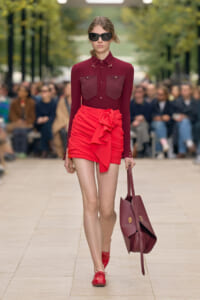 Model walking a runway in a burgundy blouse with two chest pockets and a bright red ruched mini skirt tied with a bow, paired with red shoes and a large burgundy tote, wearing dark sunglasses.