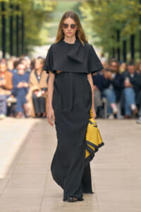 Model on a runway wearing a black draped cape dress, sunglasses, and a yellow patterned clutch/bag at her side.