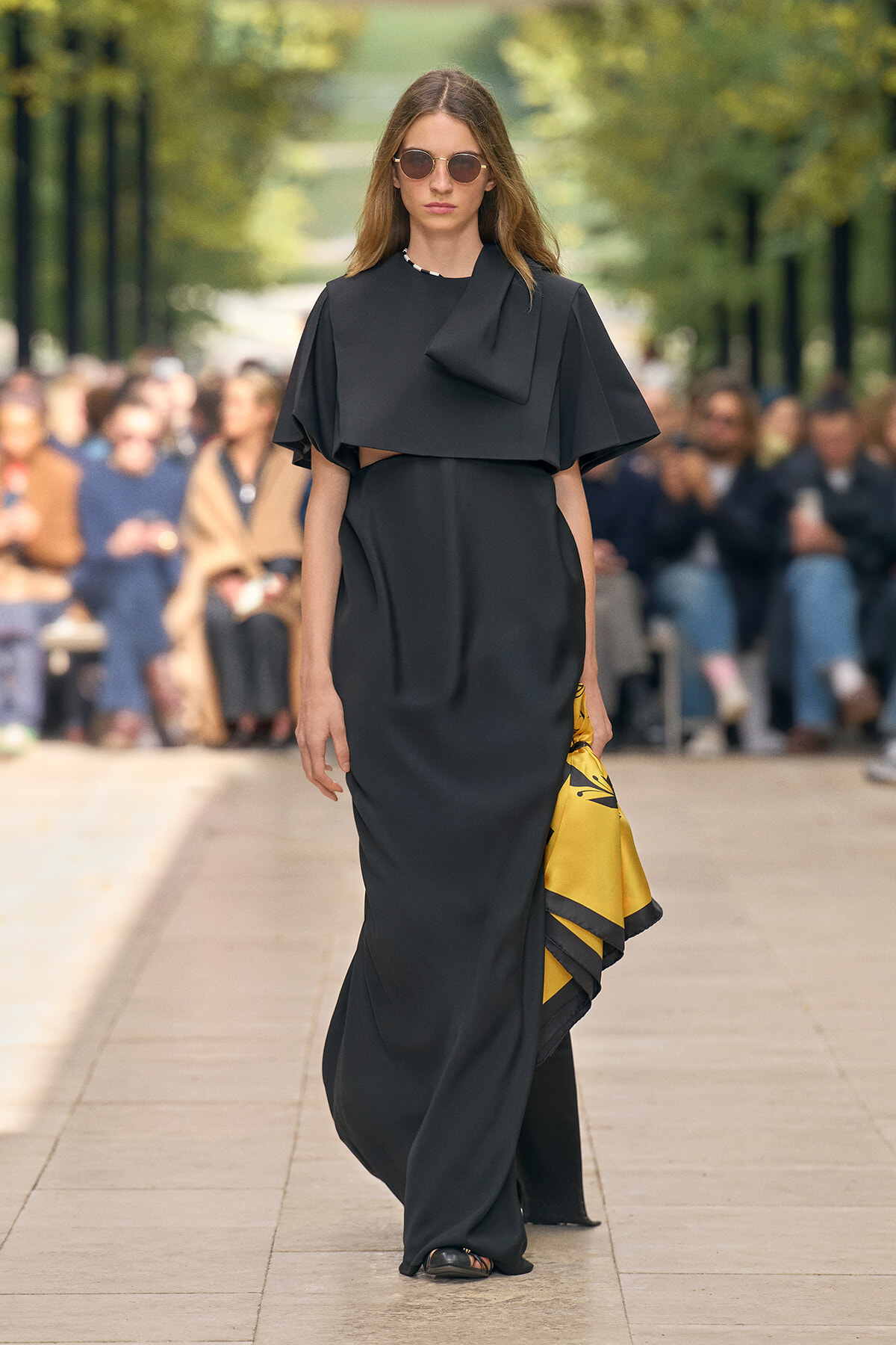 Model on a runway wearing a black draped cape dress, sunglasses, and a yellow patterned clutch/bag at her side.