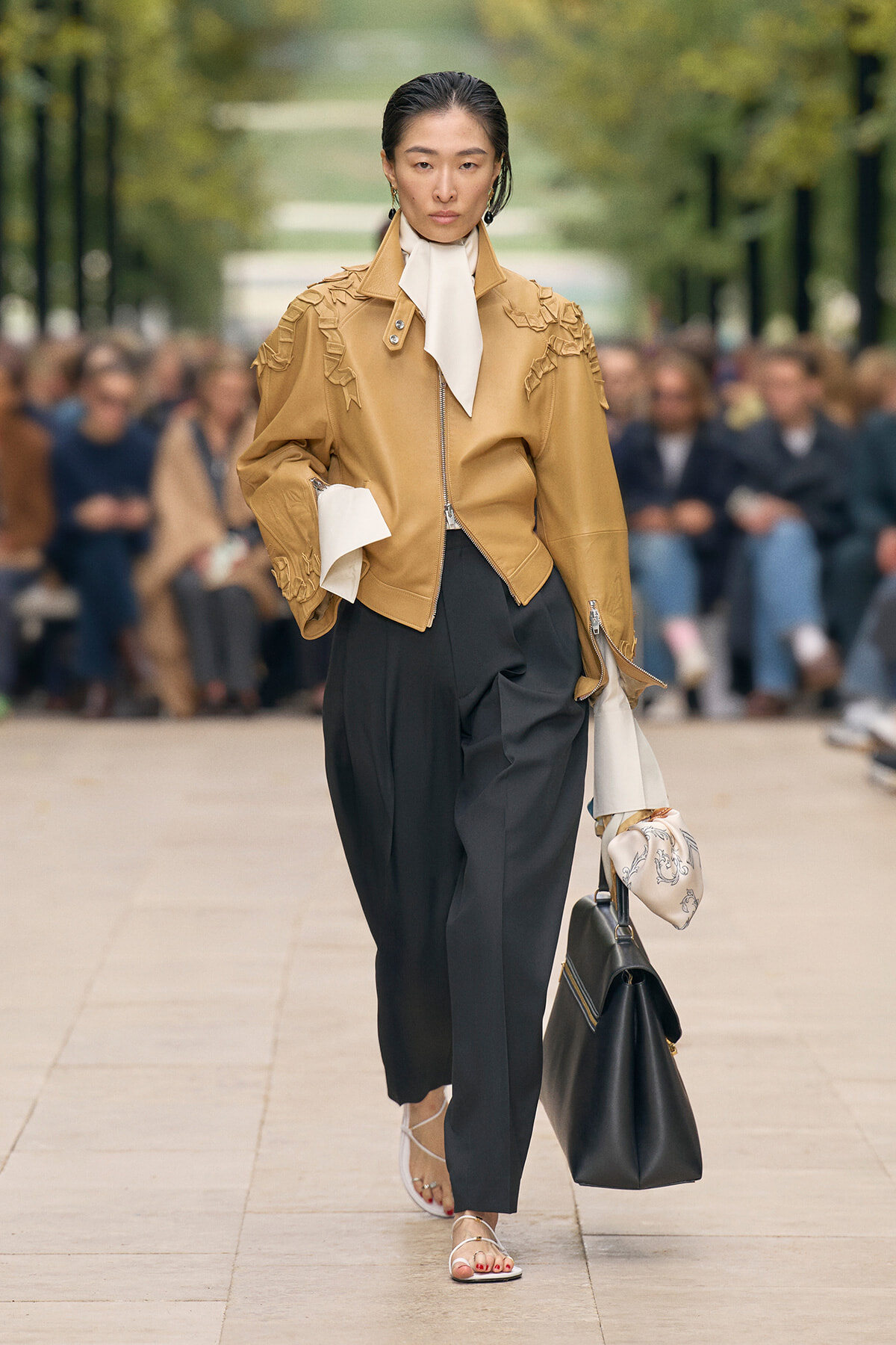 Model on a runway wearing a tan fringed leather jacket, white neck scarf, and black pleated trousers, carrying a black bag and a patterned scarf.