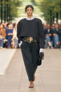Model on a runway wearing an oversized black sweater layered over a white-collared top, with wide black trousers and a metallic coin belt, carrying a small clutch.