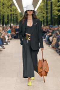 Model walks a runway in a black suit with a white bucket hat, sunglasses, and lime shoes, carrying a brown tote.