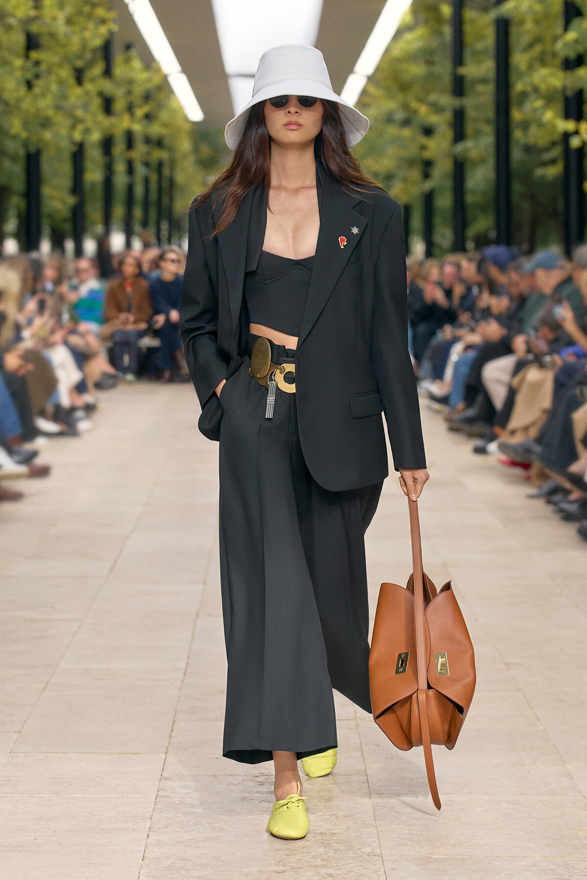 Model walks a runway in a black suit with a white bucket hat, sunglasses, and lime shoes, carrying a brown tote.