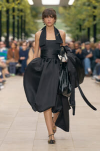 Female model walks a runway in a dramatic black halter dress with a voluminous bubble skirt, carrying a large black handbag as spectators blur in the background.