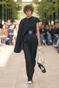 Runway model in an all-black outfit: draped cape top over one shoulder, high-waisted trousers with a wide belt, white gloves, and a light scarf bag held at the side.