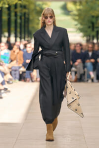 Model walking the runway in a black tailored suit with a cinched wrap belt, brown suede boots, and round sunglasses, carrying a beige printed scarf.