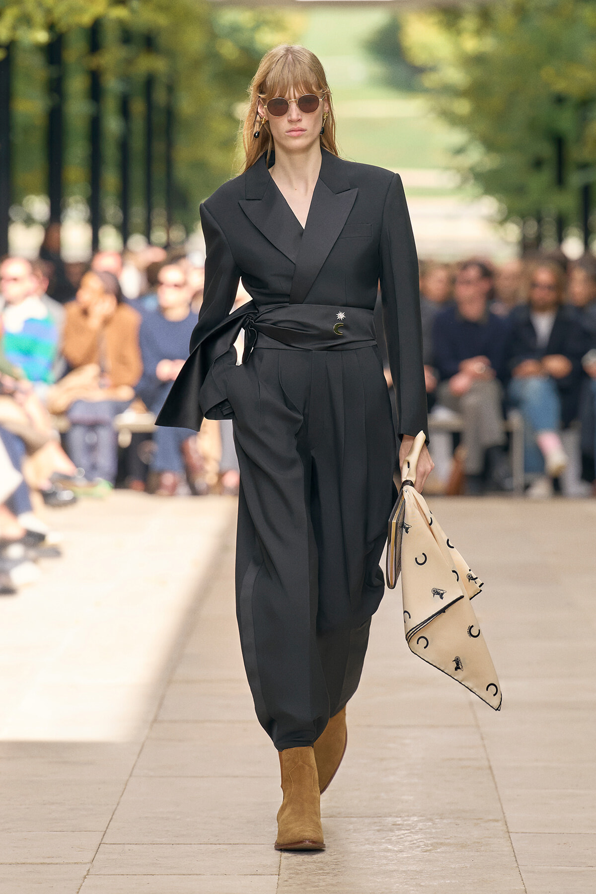 Model walking the runway in a black tailored suit with a cinched wrap belt, brown suede boots, and round sunglasses, carrying a beige printed scarf.