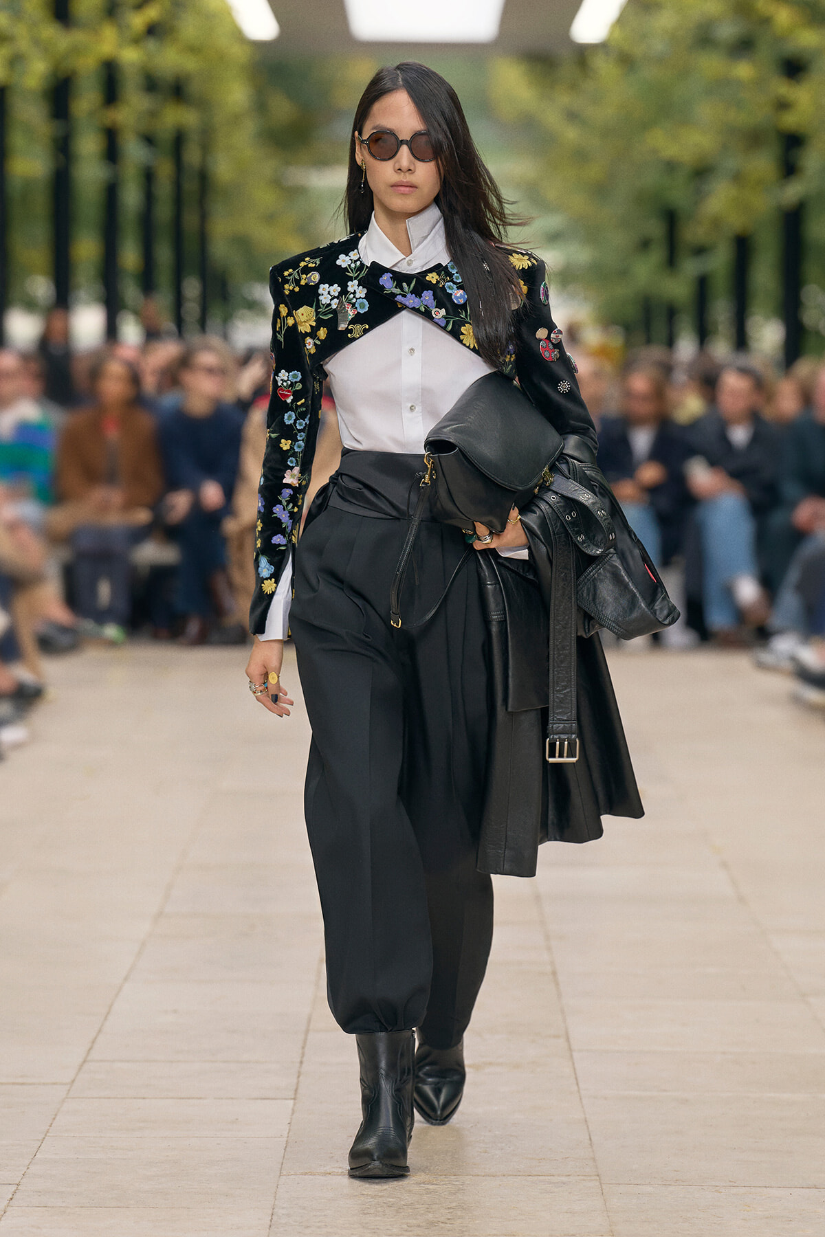 Runway model wearing a black floral-embroidered jacket over a white shirt, with loose black trousers, black boots, and a stacked black bag.