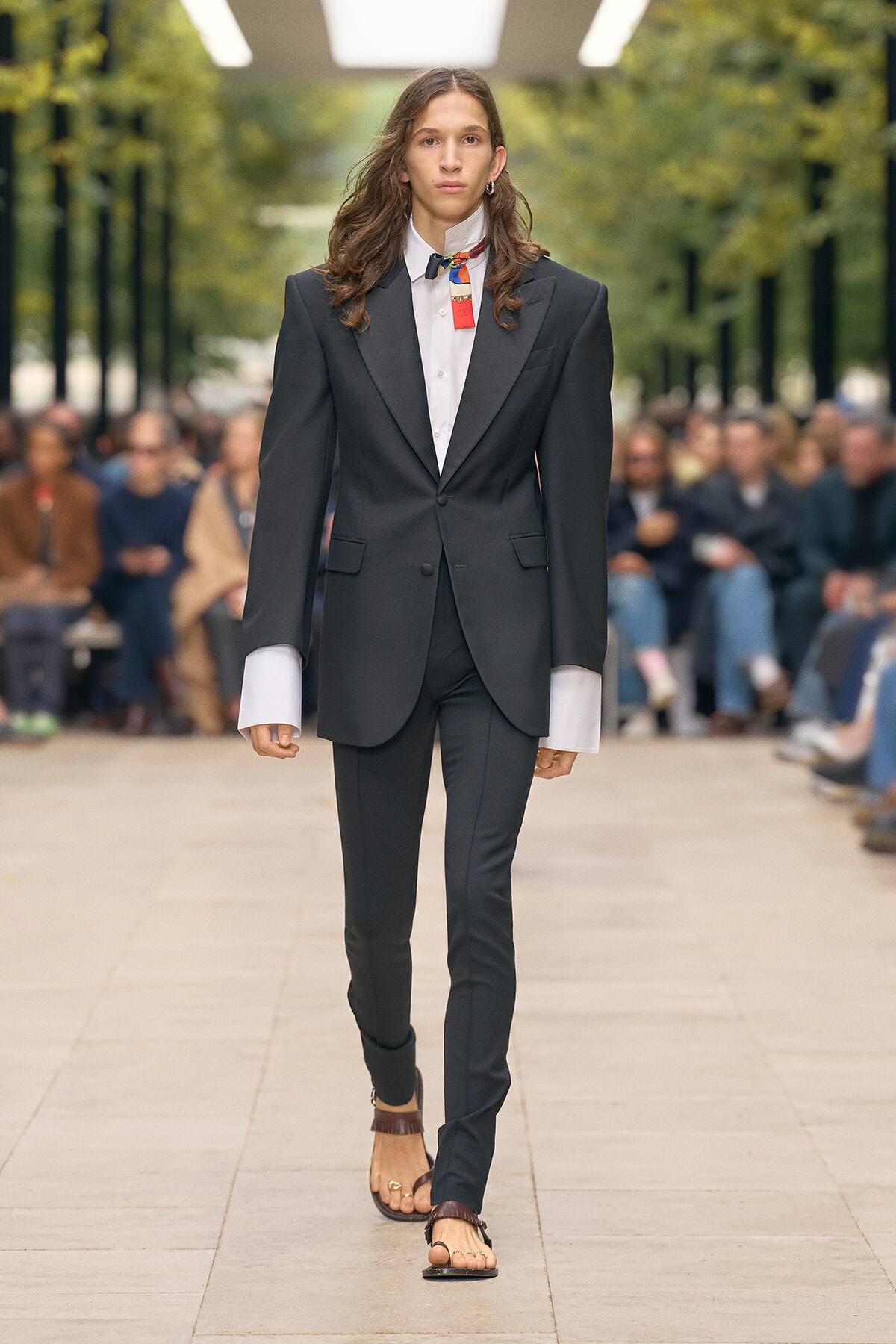 Model walks a runway in a black tailored suit with oversized white cuffed sleeves and white shirt, audience blur behind her.