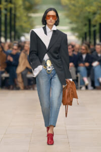 Model walking a runway in an oversized black blazer with a white lapel, light-wash jeans, red shoes, orange sunglasses, and a brown handbag.