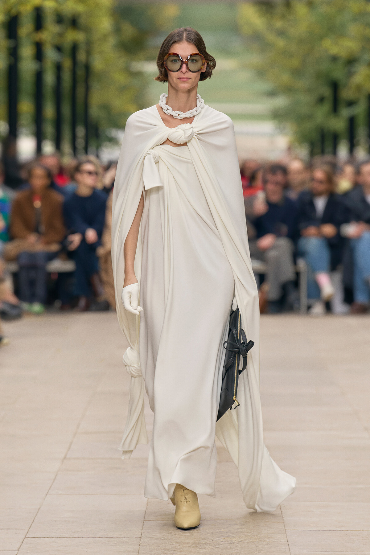 Model walks a white draped gown with oversized round sunglasses, white gloves, and a chunky necklace on a city runway. She carries a black handbag.