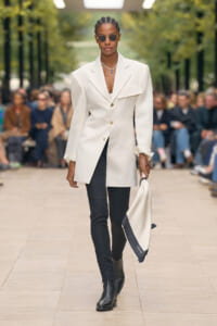 Female model walks a runway in a cream oversized blazer, black pants, and black ankle boots, wearing round sunglasses and carrying a white-and-navy scarf.