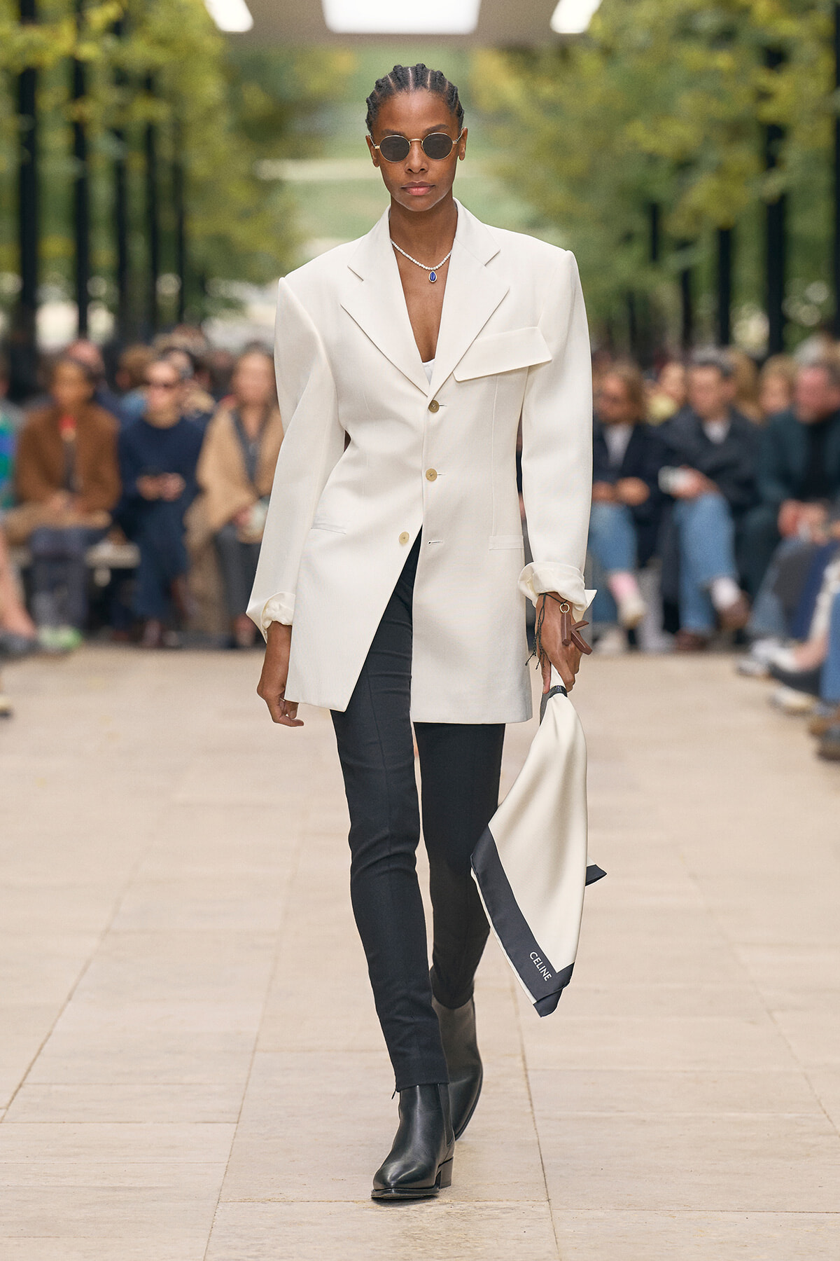 Female model walks a runway in a cream oversized blazer, black pants, and black ankle boots, wearing round sunglasses and carrying a white-and-navy scarf.