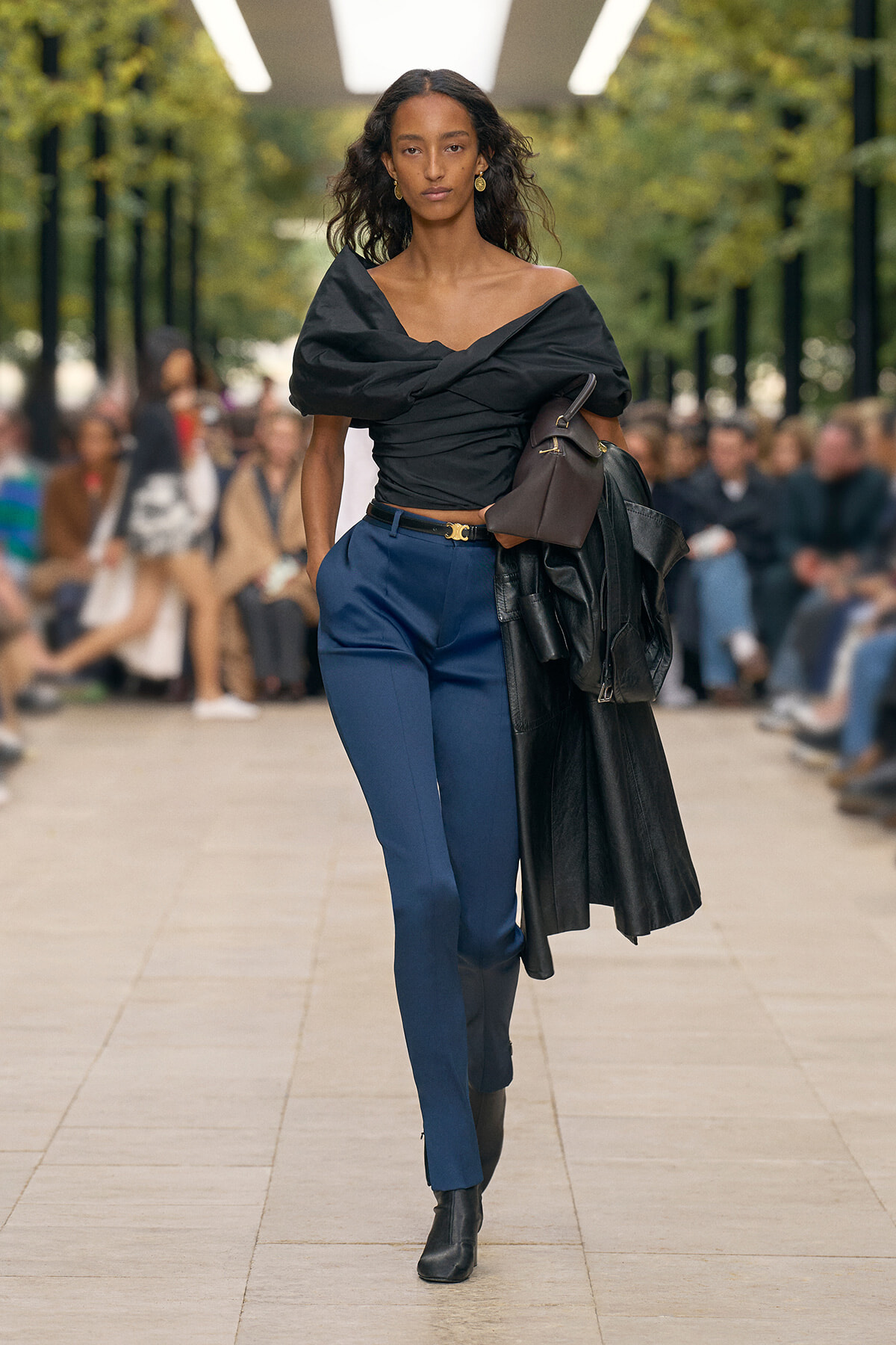 Model walking a runway outdoors in a black off‑the‑shoulder top, blue tailored pants, black ankle boots, and carrying a brown handbag with a black leather jacket draped over her arm and a belt.