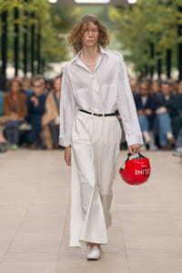 Model walks the runway in a white shirt and wide white pants, holding a red helmet at her side.