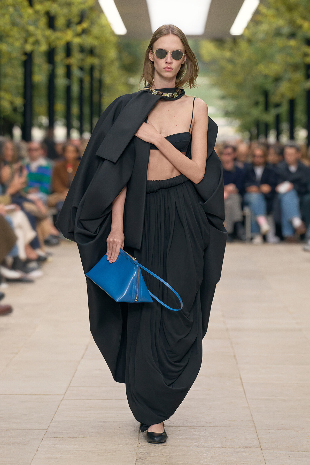 Runway model in a voluminous black draped outfit with a crop top, sunglasses, and a bright blue clutch at a fashion show capture the bold, minimal look.