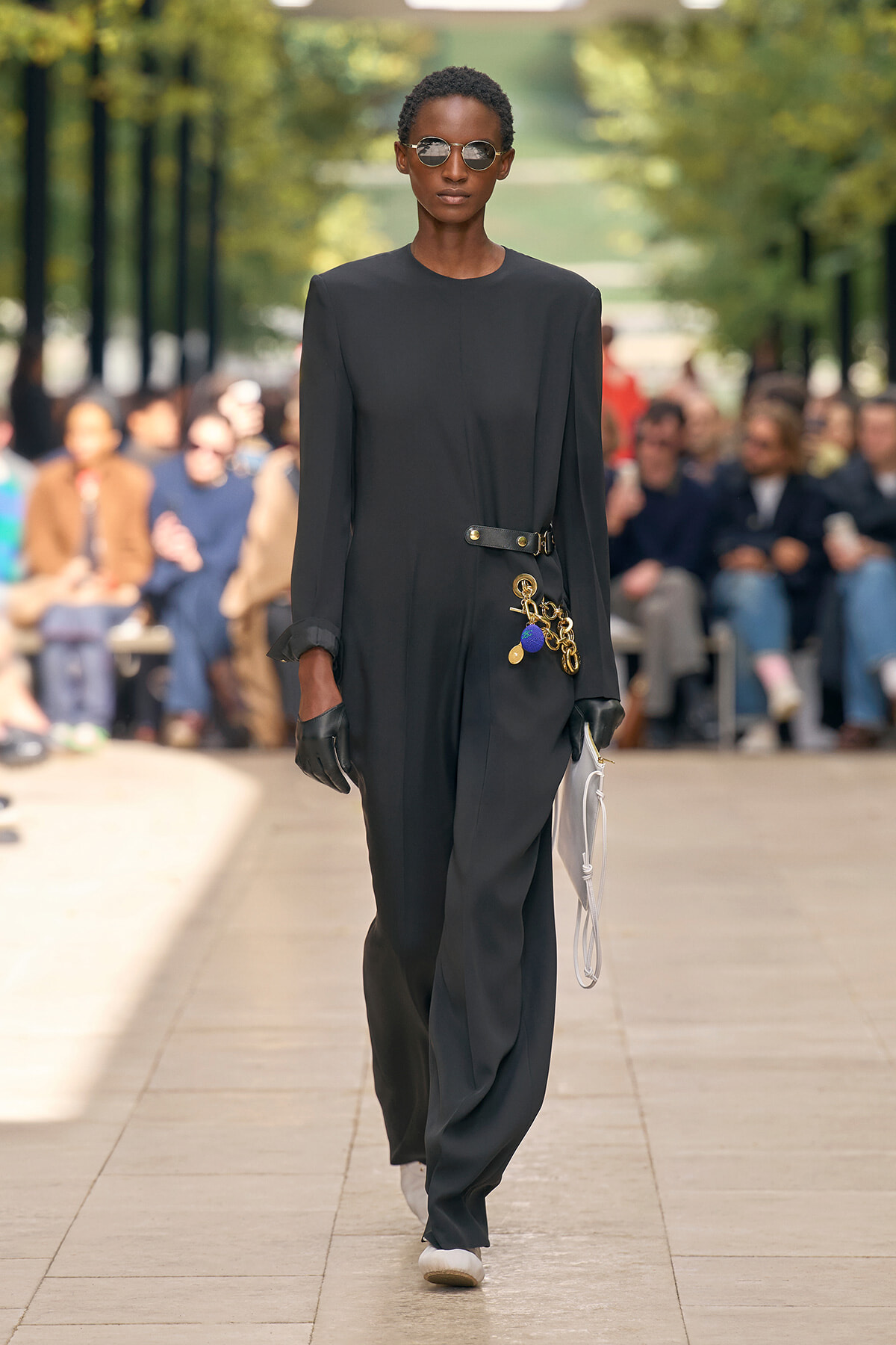 Model walks the runway in a black jumpsuit with a decorative belt and charms, wearing gloves and round sunglasses.