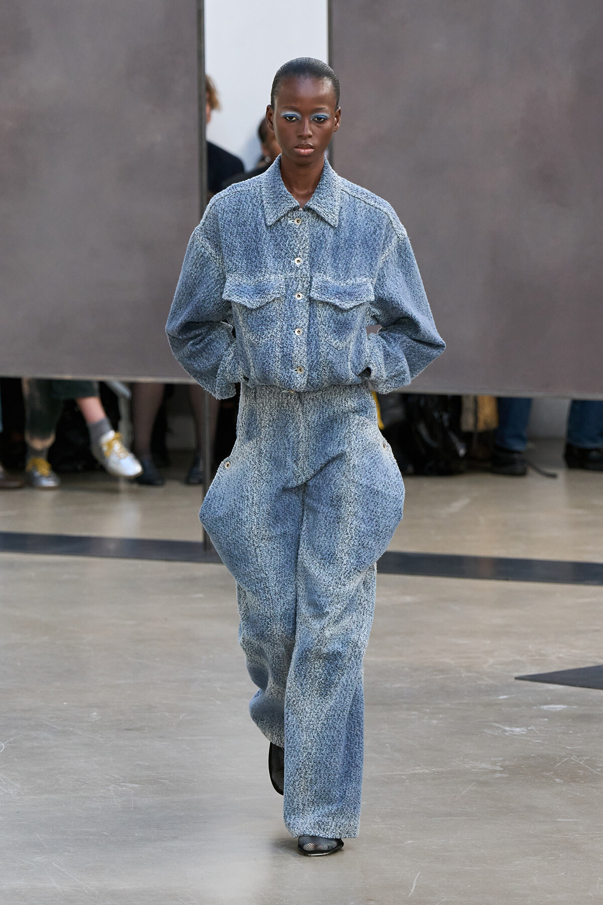 Model walks a runway wearing a matching light blue textured denim set: button-up shirt with large pockets and high-waisted wide-leg trousers, hands in pockets, neutral expression.
