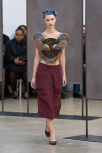 Model walks a runway in an avant-garde ensemble: sculptural heart-shaped top with fur and fabric panels, blue hair in buns, and a burgundy dotted midi skirt with heels on a neutral backdrop.