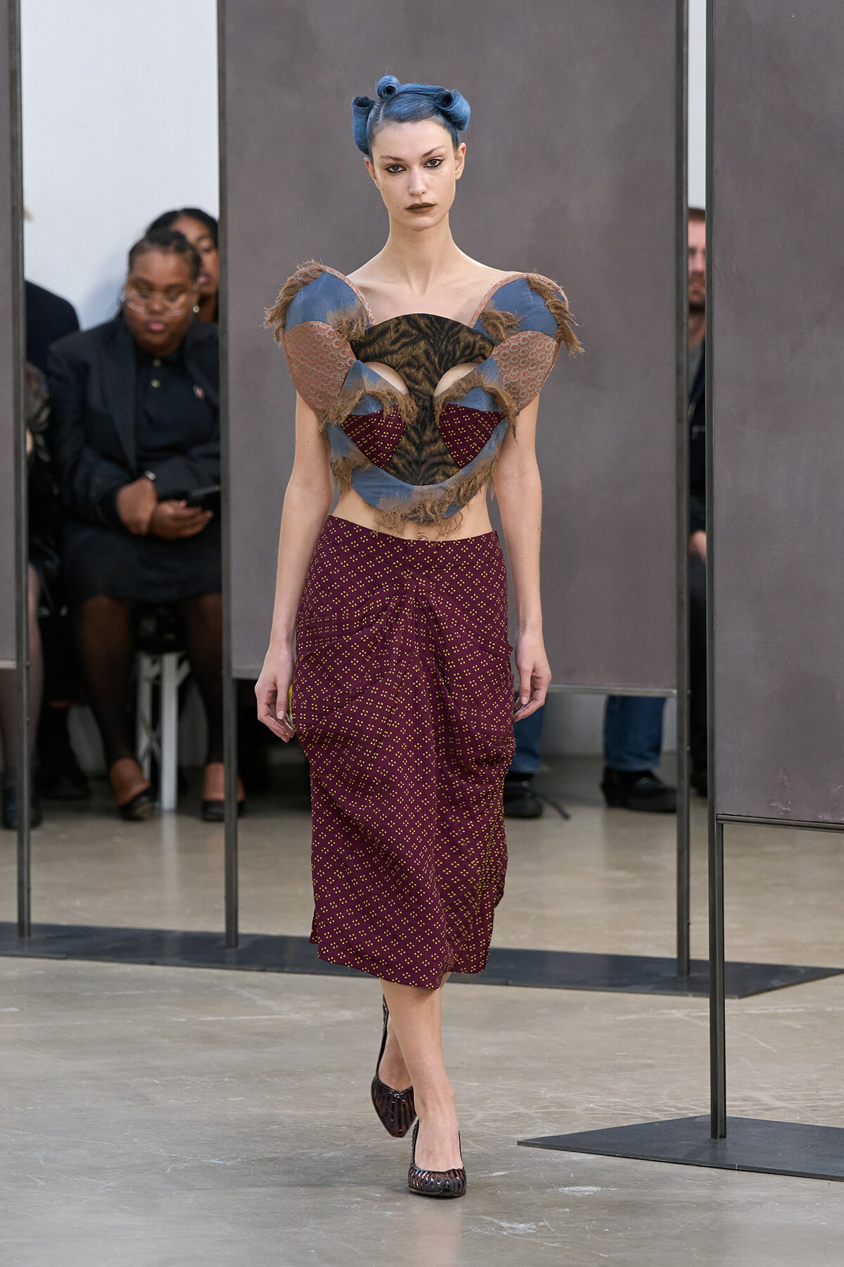 Model walks a runway in an avant-garde ensemble: sculptural heart-shaped top with fur and fabric panels, blue hair in buns, and a burgundy dotted midi skirt with heels on a neutral backdrop.