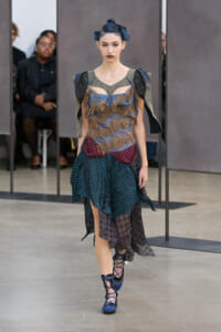 Runway model in an avant-garde ensemble: fringed gray bodice, teal skirt with burgundy panel, plaid underlayer, and blue lace-up heels.