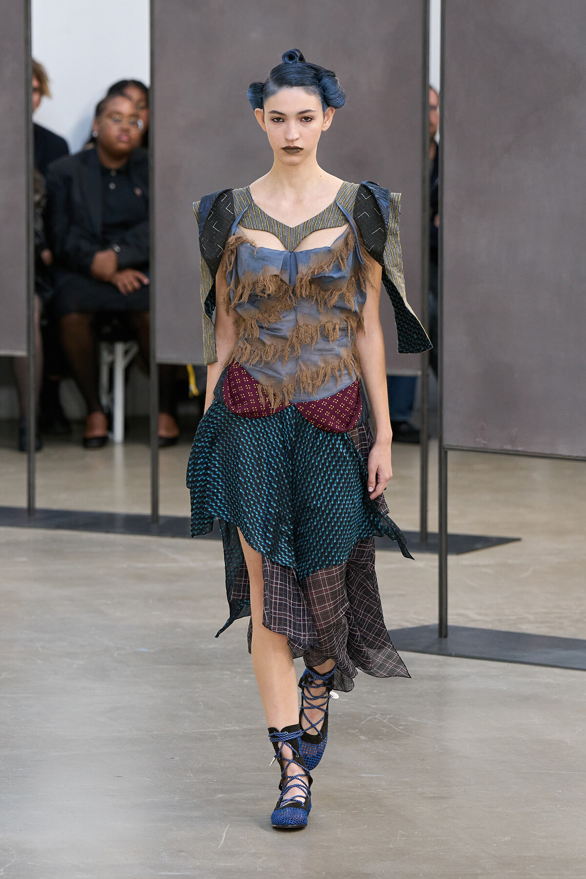 Runway model in an avant-garde ensemble: fringed gray bodice, teal skirt with burgundy panel, plaid underlayer, and blue lace-up heels.