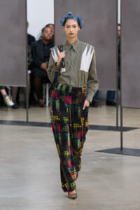Runway model in a striped shirt with white panels and colorful patchwork pants; blue hair in buns.
