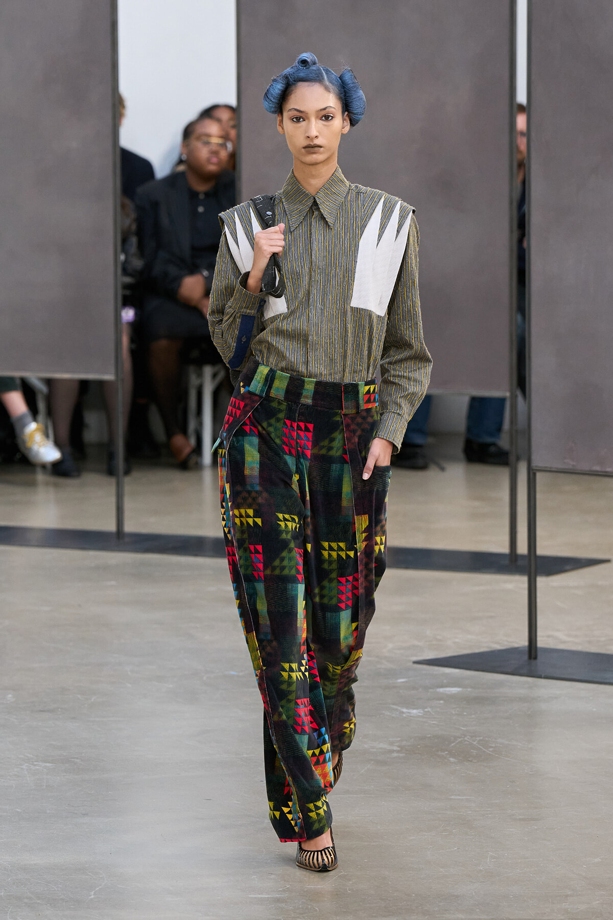 Runway model in a striped shirt with white panels and colorful patchwork pants; blue hair in buns.