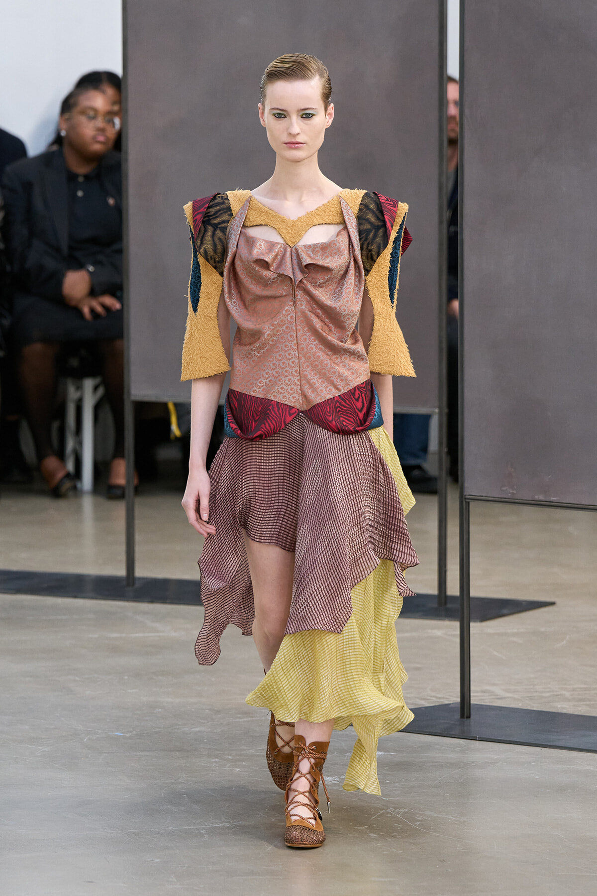 Model walks a runway in a patchwork dress with pink bustier, yellow textured sleeves, and a layered brown‑pink skirt with yellow accents, brown lace‑up sandals.