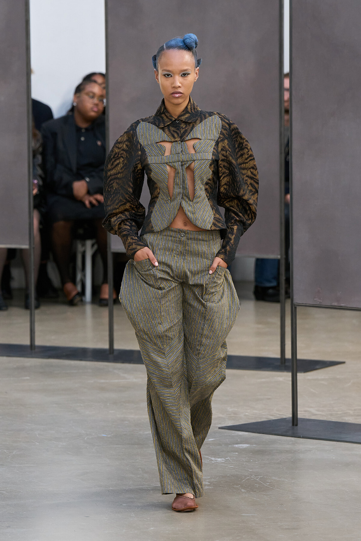 Model walks a fashion runway in a structured gray-green pinstripe two-piece with cutouts and brown patterned puff sleeves, blue bun hairstyle.