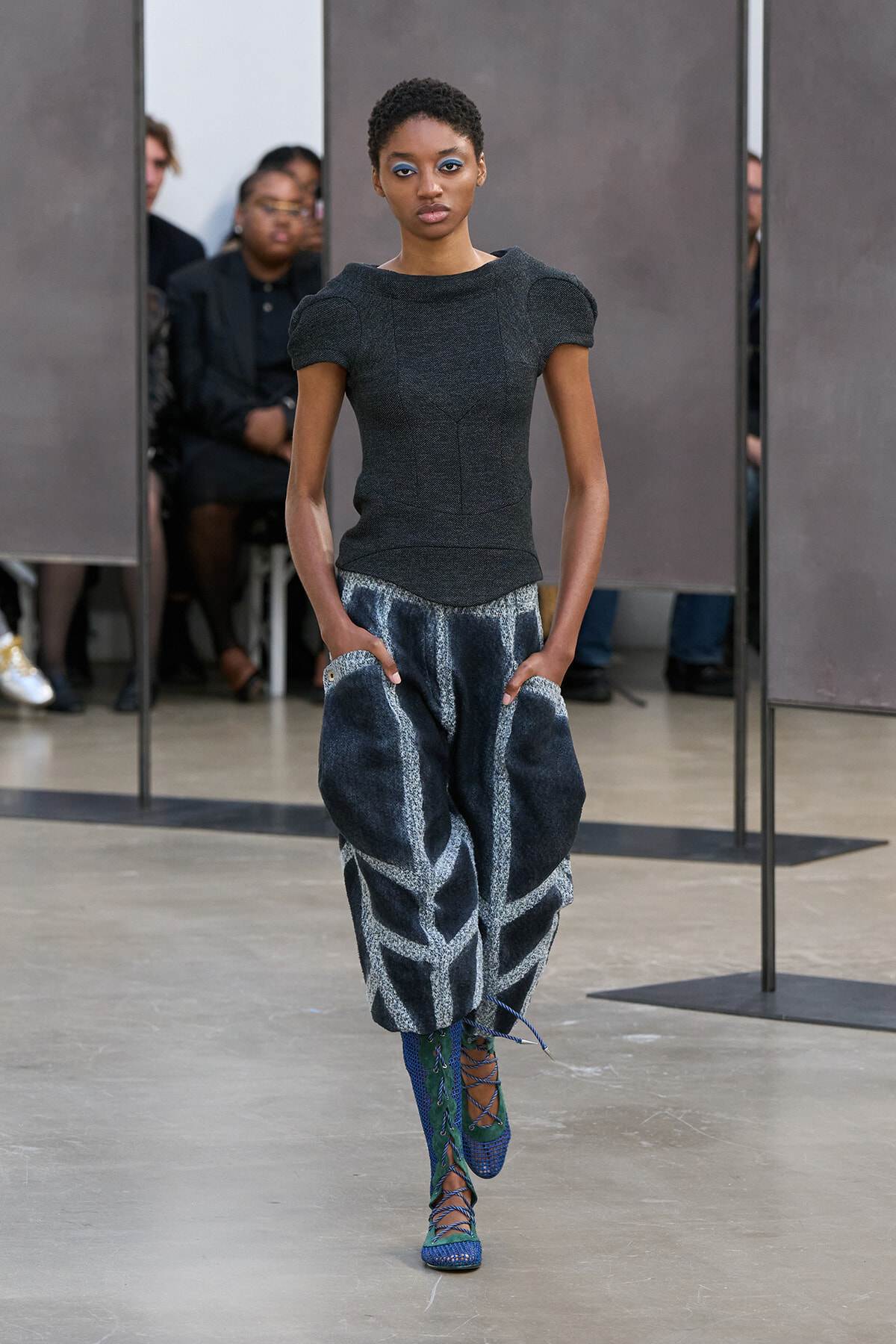 Model walking a runway in a fitted dark knit top with puff sleeves and oversized abstract-patterned pants, turquoise lace-up boots visible behind.