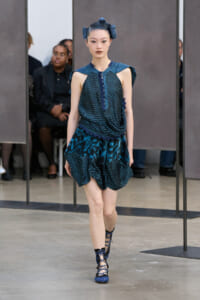 Model walking the runway in a blue-green patterned sleeveless top and ruffled skirt with lace-up sandals.